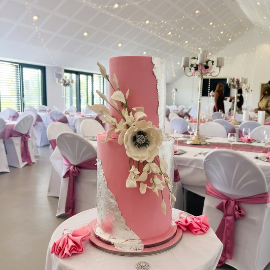 Pink wedding cake with floral decorations on a table in a decorated room.